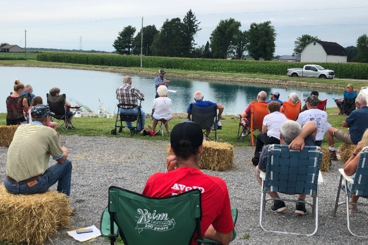 Group of people seated on hay bales and lawn chairs along a pond, listening to a man speaking at an outdoor gathering on a rural, sunny day.