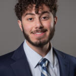 Smiling man in a navy suit and striped tie poses for a professional headshot against a gray backdrop, facing the camera.