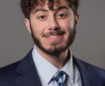Smiling man in a navy suit and striped tie poses for a professional headshot against a gray backdrop, facing the camera.