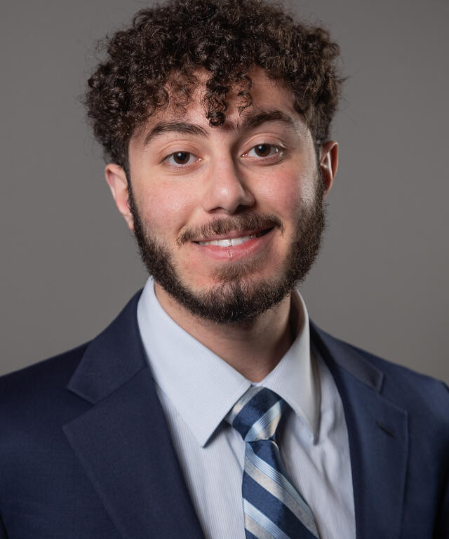 Smiling man in a navy suit and striped tie poses for a professional headshot against a gray backdrop, facing the camera.