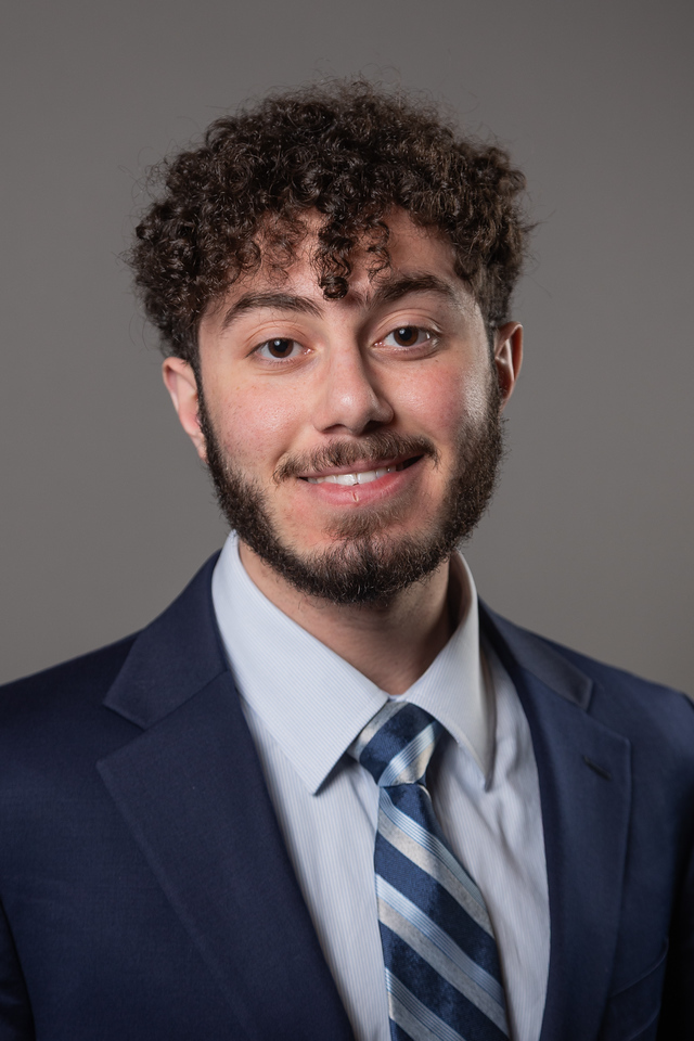 Smiling man in a navy suit and striped tie poses for a professional headshot against a gray backdrop, facing the camera.