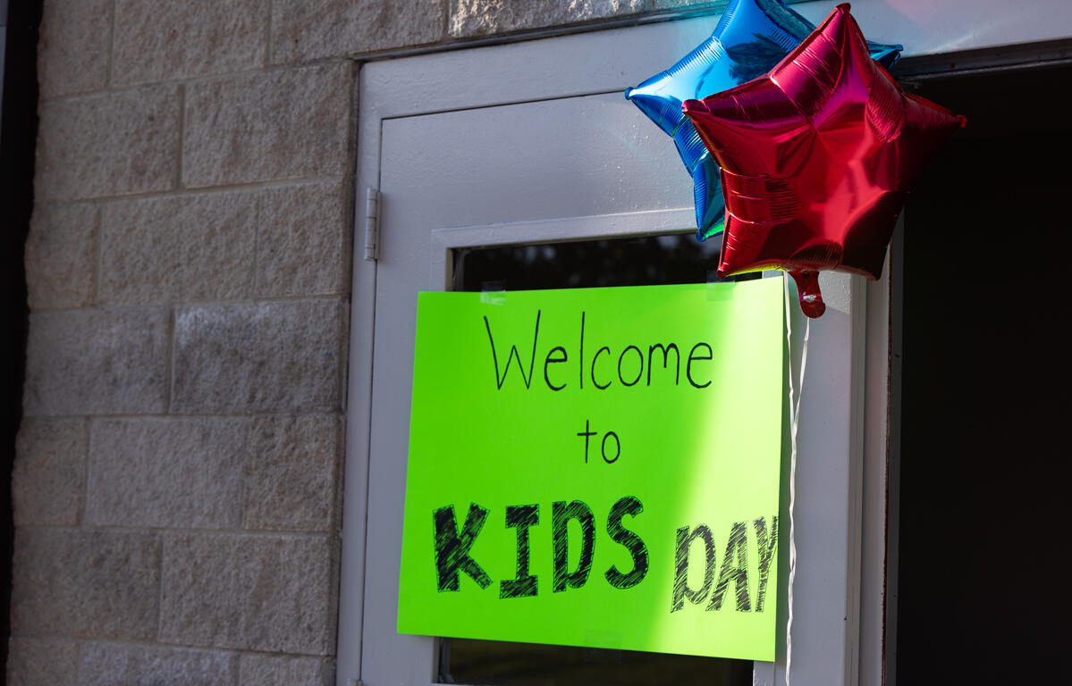 Bright neon green sign on a door that reads 'Welcome to KIDS day', with blue and red star balloons above.