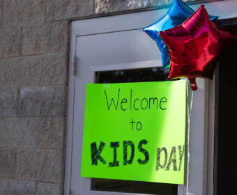 Bright neon green sign on a door that reads 'Welcome to KIDS day', with blue and red star balloons above.