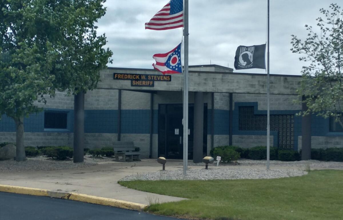 Front of a sheriff's office with three flagpoles: a U.S. flag, a red-white-blue state flag, and a third banner flag, plus a sign reading 'FREDRICK W. STEVENS SHERIFF' on the building façade.