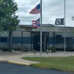 Front of a sheriff's office with three flagpoles: a U.S. flag, a red-white-blue state flag, and a third banner flag, plus a sign reading 'FREDRICK W. STEVENS SHERIFF' on the building façade.