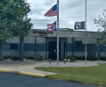 Front of a sheriff's office with three flagpoles: a U.S. flag, a red-white-blue state flag, and a third banner flag, plus a sign reading 'FREDRICK W. STEVENS SHERIFF' on the building façade.