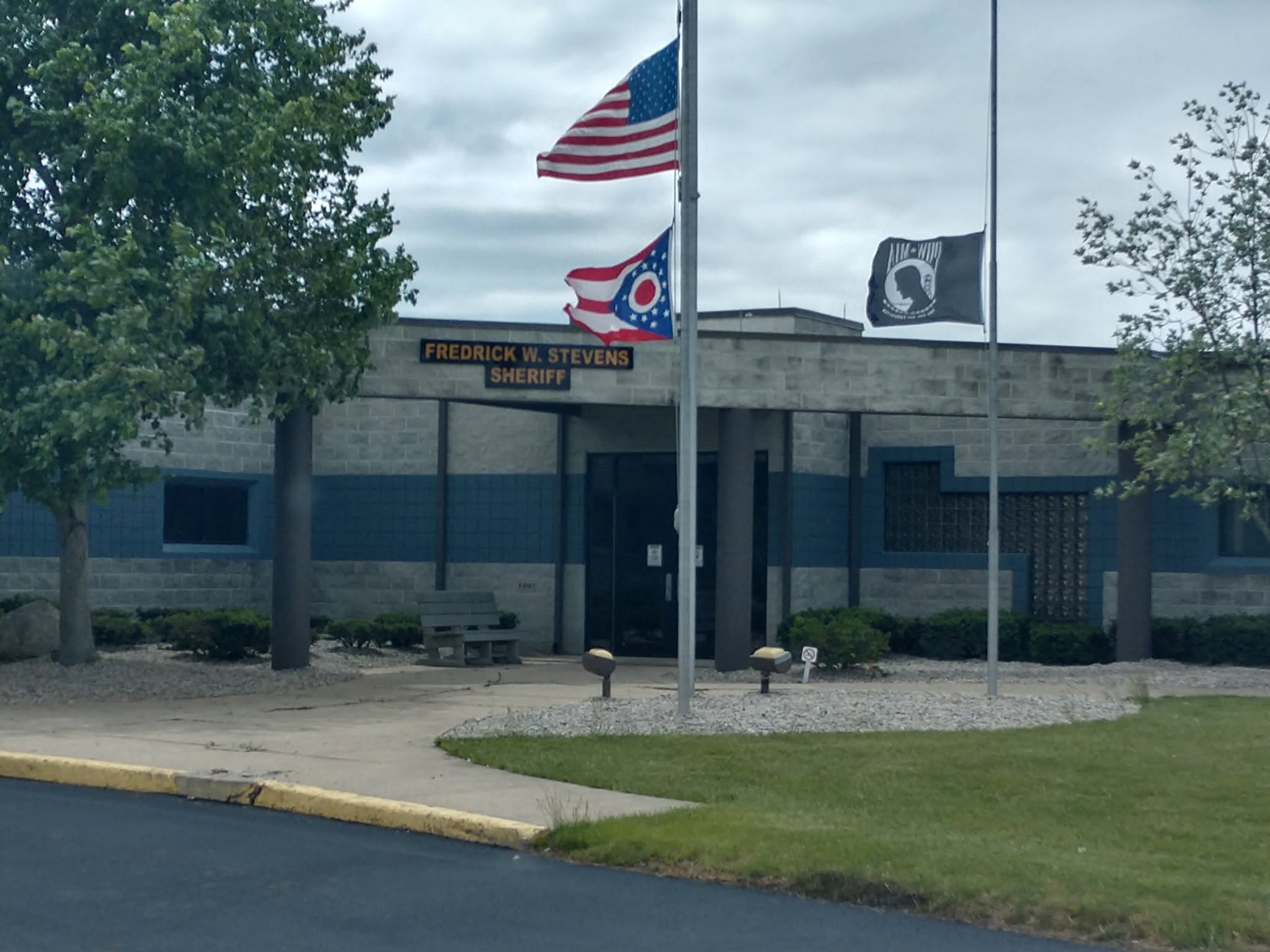 Front of a sheriff's office with three flagpoles: a U.S. flag, a red-white-blue state flag, and a third banner flag, plus a sign reading 'FREDRICK W. STEVENS SHERIFF' on the building façade.