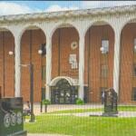 Stone-faced government building with a row of arches and a manicured lawn; in the foreground a large cannon and a black memorial wall with inscriptions.