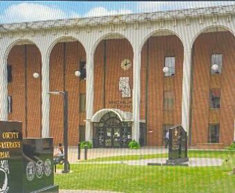 Stone-faced government building with a row of arches and a manicured lawn; in the foreground a large cannon and a black memorial wall with inscriptions.