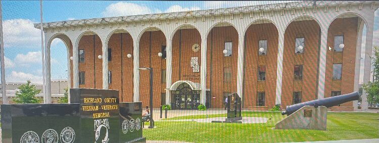Stone-faced government building with a row of arches and a manicured lawn; in the foreground a large cannon and a black memorial wall with inscriptions.