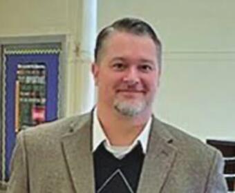Man in a beige blazer and white shirt standing indoors, smiling at the camera.