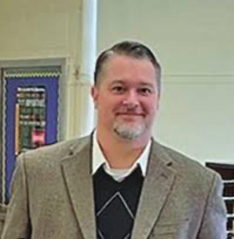 Man in a beige blazer and white shirt standing indoors, smiling at the camera.