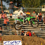 Motocross rider on a green bike launches over a dirt jump as spectators watch from the bleachers in the background.