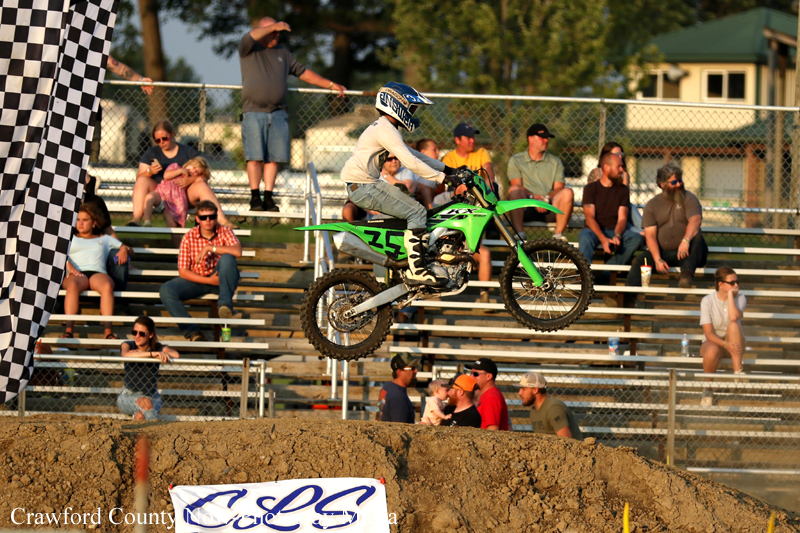 Motocross rider on a green bike launches over a dirt jump as spectators watch from the bleachers in the background.