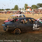 Demolition derby car #26, a black sedan, crashes on a muddy dirt track with other damaged cars nearby and spectators in the stands behind.