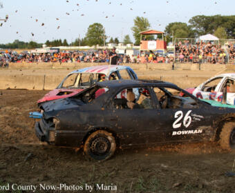 Demolition derby car #26, a black sedan, crashes on a muddy dirt track with other damaged cars nearby and spectators in the stands behind.