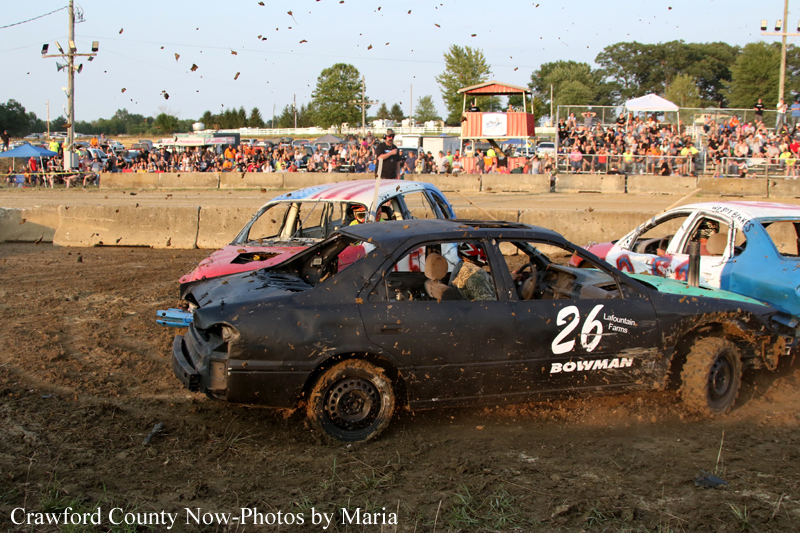 Demolition derby car #26, a black sedan, crashes on a muddy dirt track with other damaged cars nearby and spectators in the stands behind.