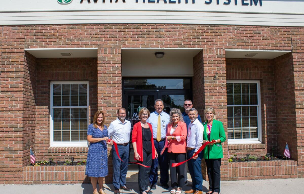 Eight people at a ribbon-cutting ceremony in front of an Avita Health System brick building, holding a red ribbon across the doorway.
