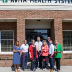 Eight people at a ribbon-cutting ceremony in front of an Avita Health System brick building, holding a red ribbon across the doorway.