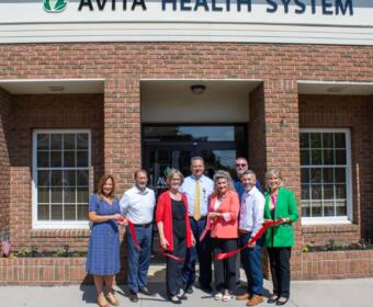 Eight people at a ribbon-cutting ceremony in front of an Avita Health System brick building, holding a red ribbon across the doorway.