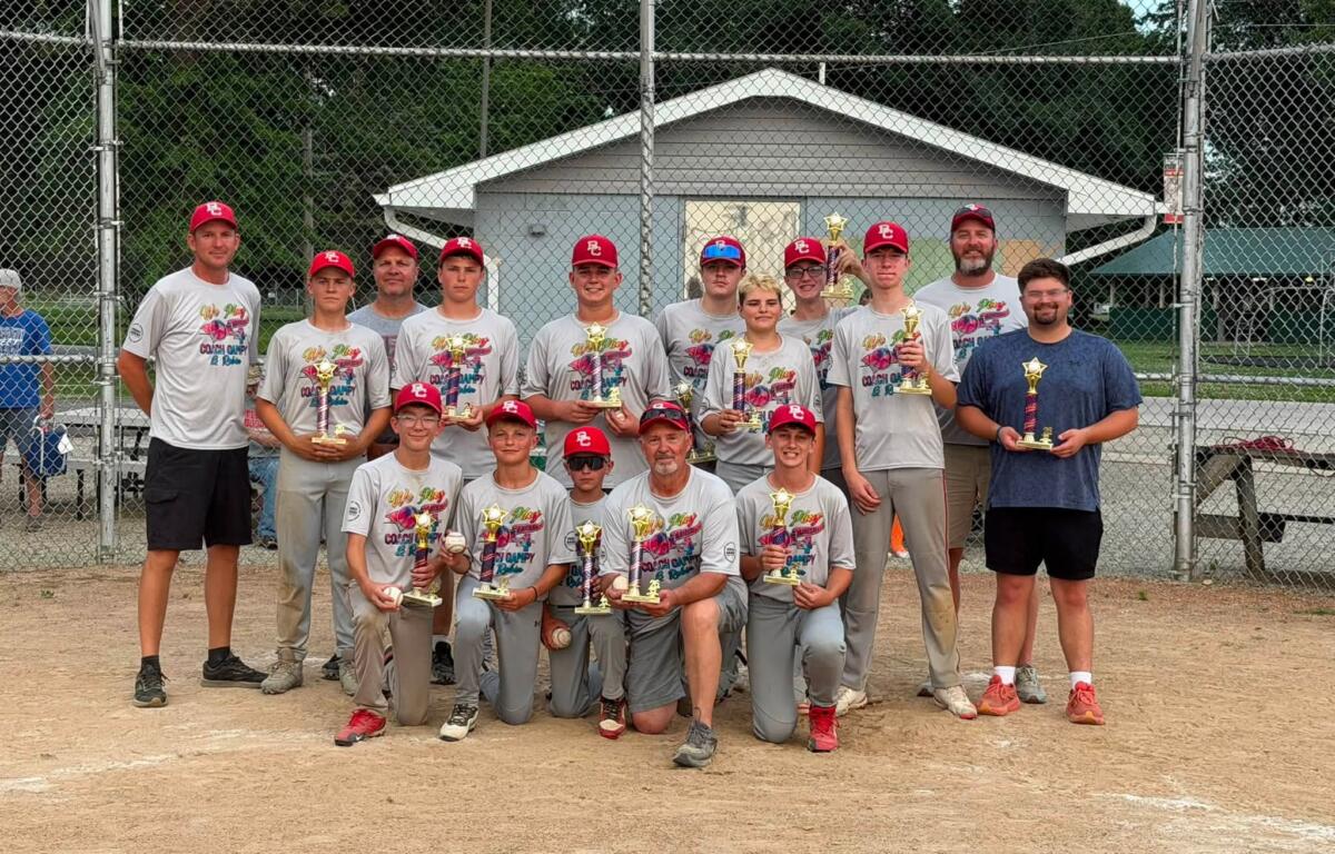 Youth baseball team posing with trophies in front of a chain-link fence after a game, wearing gray shirts and red caps.