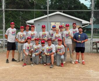 Youth baseball team posing with trophies in front of a chain-link fence after a game, wearing gray shirts and red caps.
