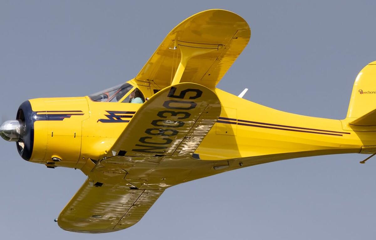 Bright yellow vintage single-engine airplane in flight, blue nose, cockpit visible against a clear blue sky, registration on the wing.
