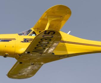 Bright yellow vintage single-engine airplane in flight, blue nose, cockpit visible against a clear blue sky, registration on the wing.