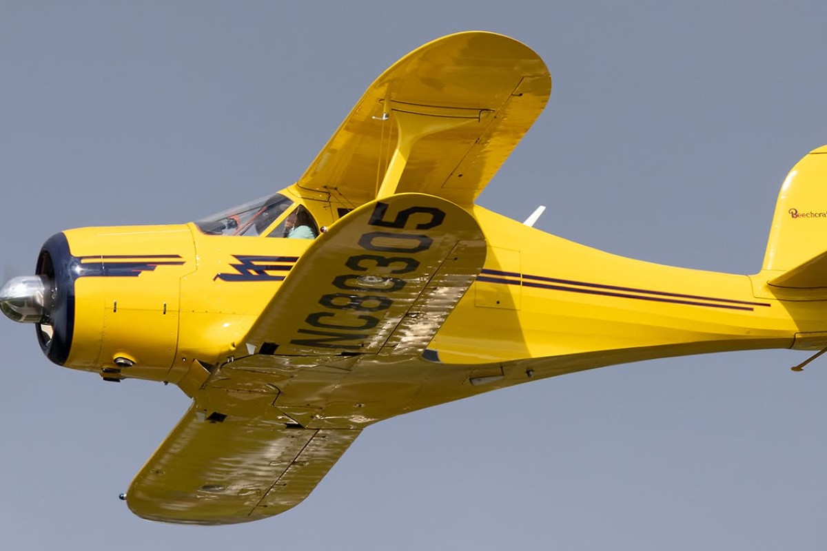 Bright yellow vintage single-engine airplane in flight, blue nose, cockpit visible against a clear blue sky, registration on the wing.