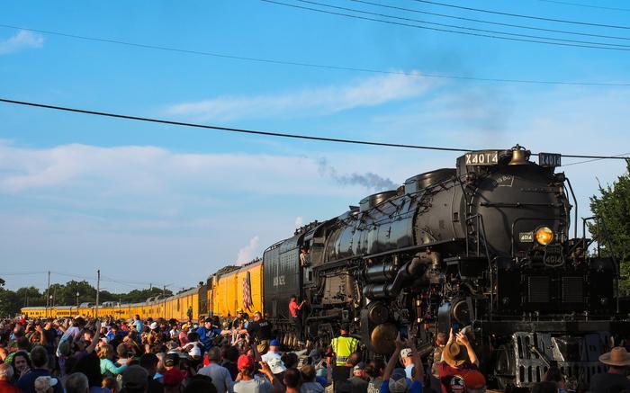 Steam locomotive pulling yellow passenger cars on a sunny day as a crowd gathers along the tracks for a train event.