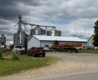 Farmyard with metal grain silos beside a white barn; there are vehicles and a boat on a trailer in the foreground under a cloudy sky.