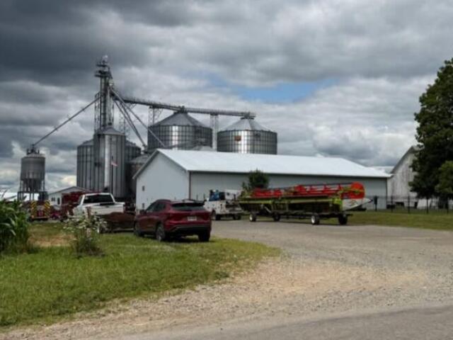 Farmyard with metal grain silos beside a white barn; there are vehicles and a boat on a trailer in the foreground under a cloudy sky.