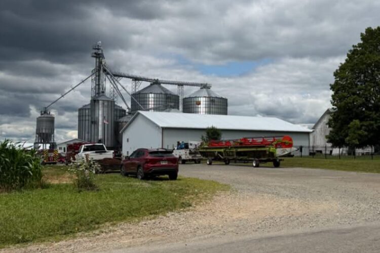 Farmyard with metal grain silos beside a white barn; there are vehicles and a boat on a trailer in the foreground under a cloudy sky.