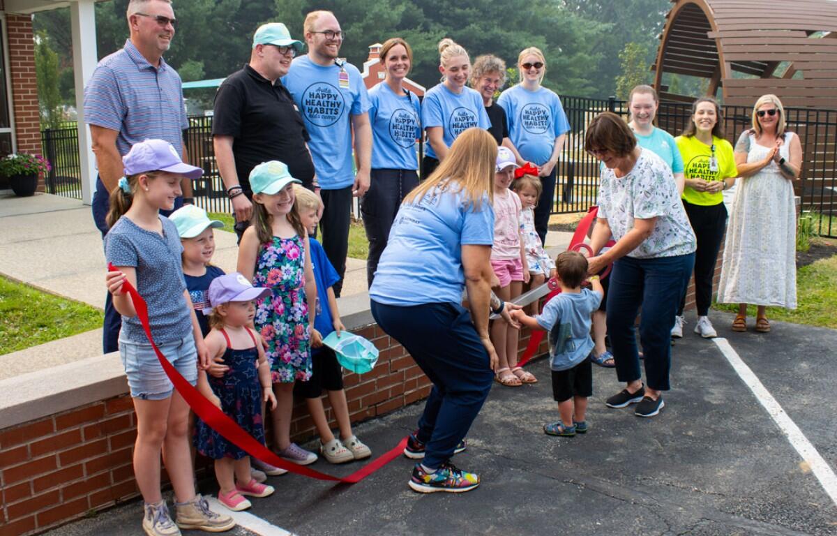 Crowd of adults and children at an outdoor ribbon-cutting ceremony; a woman hands a toddler the red ribbon while others watch, wearing light-blue shirts and hats.