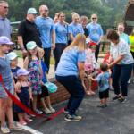 Crowd of adults and children at an outdoor ribbon-cutting ceremony; a woman hands a toddler the red ribbon while others watch, wearing light-blue shirts and hats.