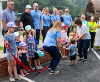 Crowd of adults and children at an outdoor ribbon-cutting ceremony; a woman hands a toddler the red ribbon while others watch, wearing light-blue shirts and hats.