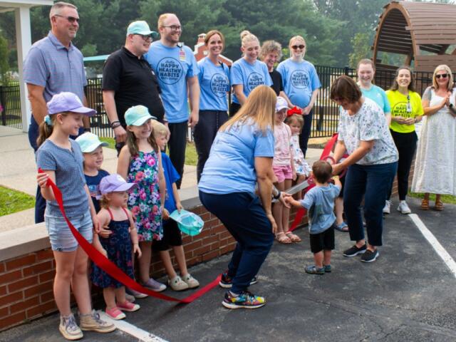 Crowd of adults and children at an outdoor ribbon-cutting ceremony; a woman hands a toddler the red ribbon while others watch, wearing light-blue shirts and hats.
