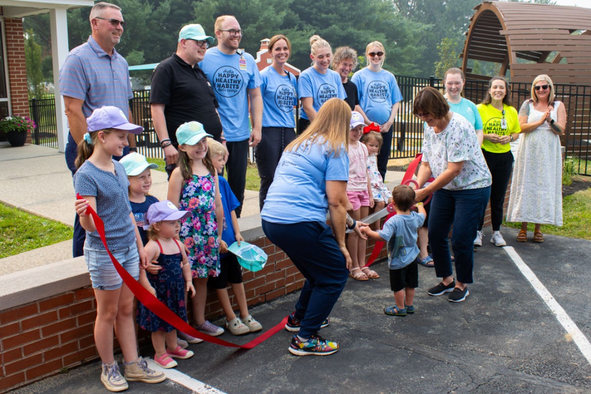 Crowd of adults and children at an outdoor ribbon-cutting ceremony; a woman hands a toddler the red ribbon while others watch, wearing light-blue shirts and hats.
