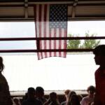 American flag hanging from a ceiling, center of the frame, above a darkened crowd at a covered outdoor venue.