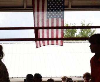 American flag hanging from a ceiling, center of the frame, above a darkened crowd at a covered outdoor venue.