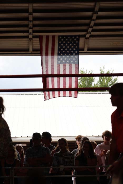 American flag hanging from a ceiling, center of the frame, above a darkened crowd at a covered outdoor venue.