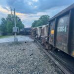 Derailed freight train with mangled cars on the tracks beside a crossing signal at dusk.