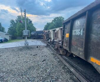 Derailed freight train with mangled cars on the tracks beside a crossing signal at dusk.
