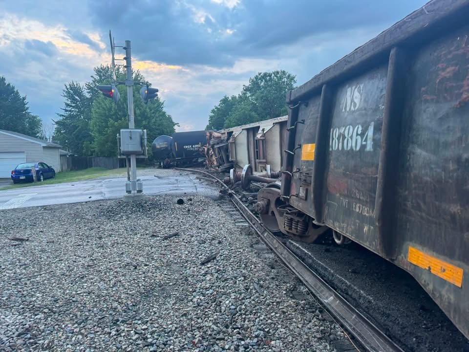 Derailed freight train with mangled cars on the tracks beside a crossing signal at dusk.