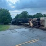 Derailed freight train with a toppled tanker car on tracks beside a road, trees in the background.