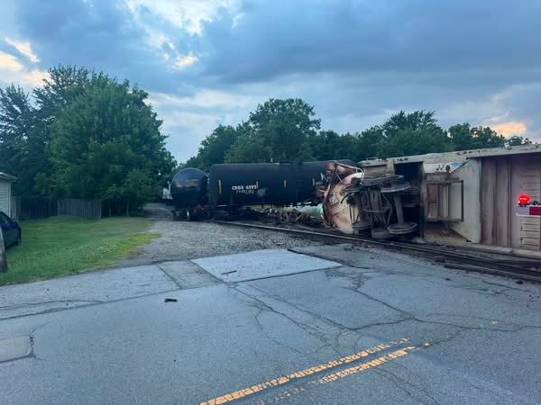 Derailed freight train with a toppled tanker car on tracks beside a road, trees in the background.