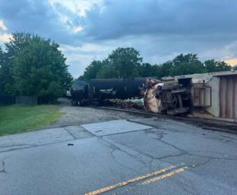 Derailed train cars on tracks beside a rural road, including a tilted tanker and overturned freight car with trees in the background.