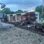 Derailed freight car on its side across the tracks with scattered debris; crossing signals and damaged wheelsets nearby, in a rural-suburban setting with trees in the background.