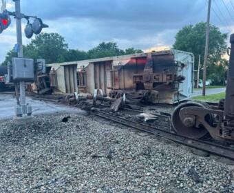Derailed freight car on its side across the tracks with scattered debris; crossing signals and damaged wheelsets nearby, in a rural-suburban setting with trees in the background.