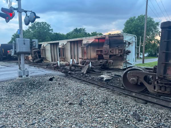 Derailed freight car on its side across the tracks with scattered debris; crossing signals and damaged wheelsets nearby, in a rural-suburban setting with trees in the background.